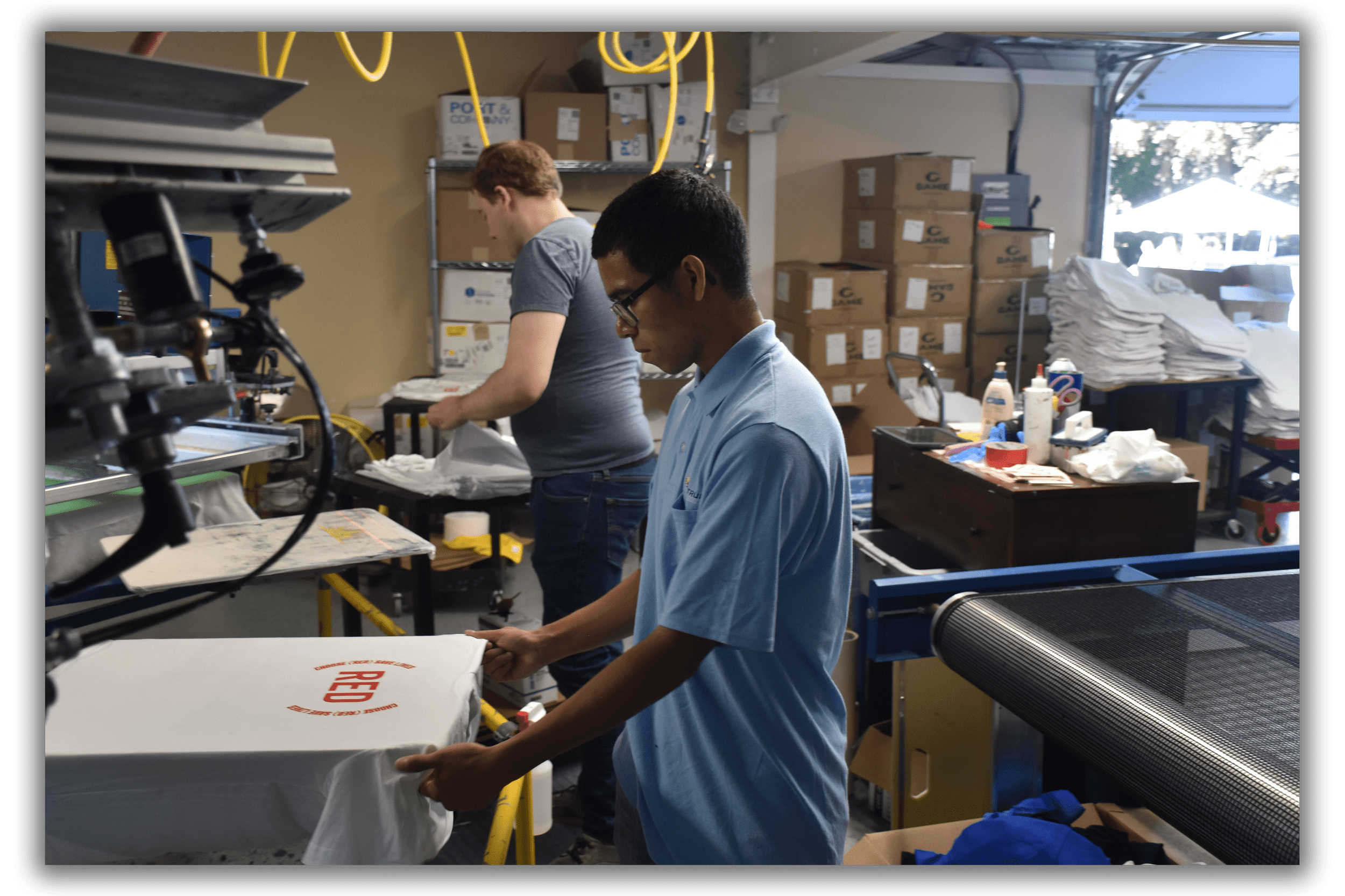 Two people working in a t-shirt printing facility, handling shirts near printing and drying equipment. Boxes and supplies are stacked in the background.